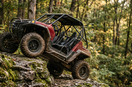 Dark red side-by-side climbing a mossy rock ledge in a hardwood forest, skid plate and A-arm guards visible below